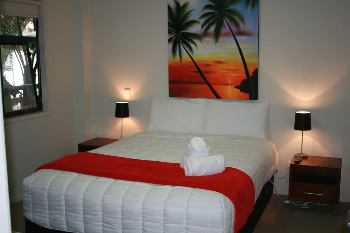 A neatly made bed with a red blanket, two bedside lamps, and a painting of a sunset with palm trees on the wall.