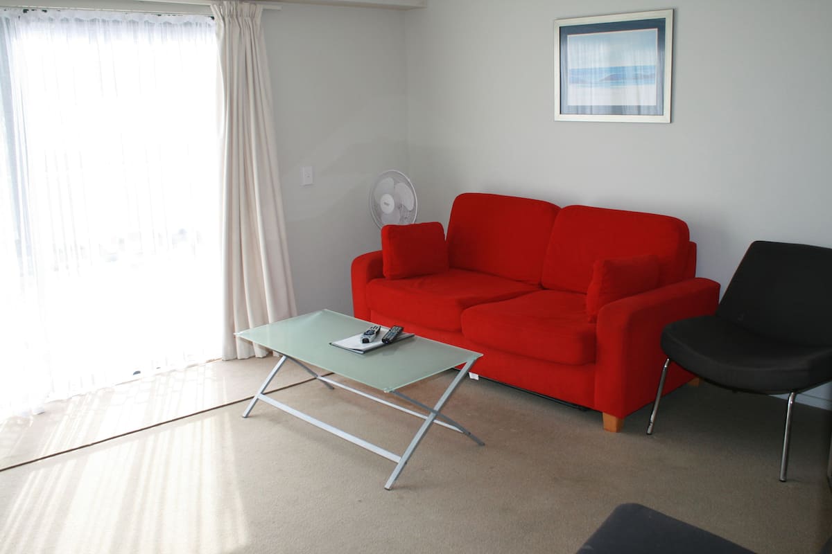 A living room with a red sofa, a black chair, a green coffee table, and a standing fan.