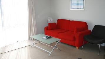 Un salon avec un canapé rouge, un fauteuil noir, une table basse verte et un ventilateur sur pied.