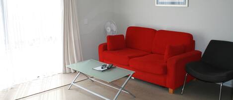 A living room with a red sofa, a black chair, a green coffee table, and a standing fan.