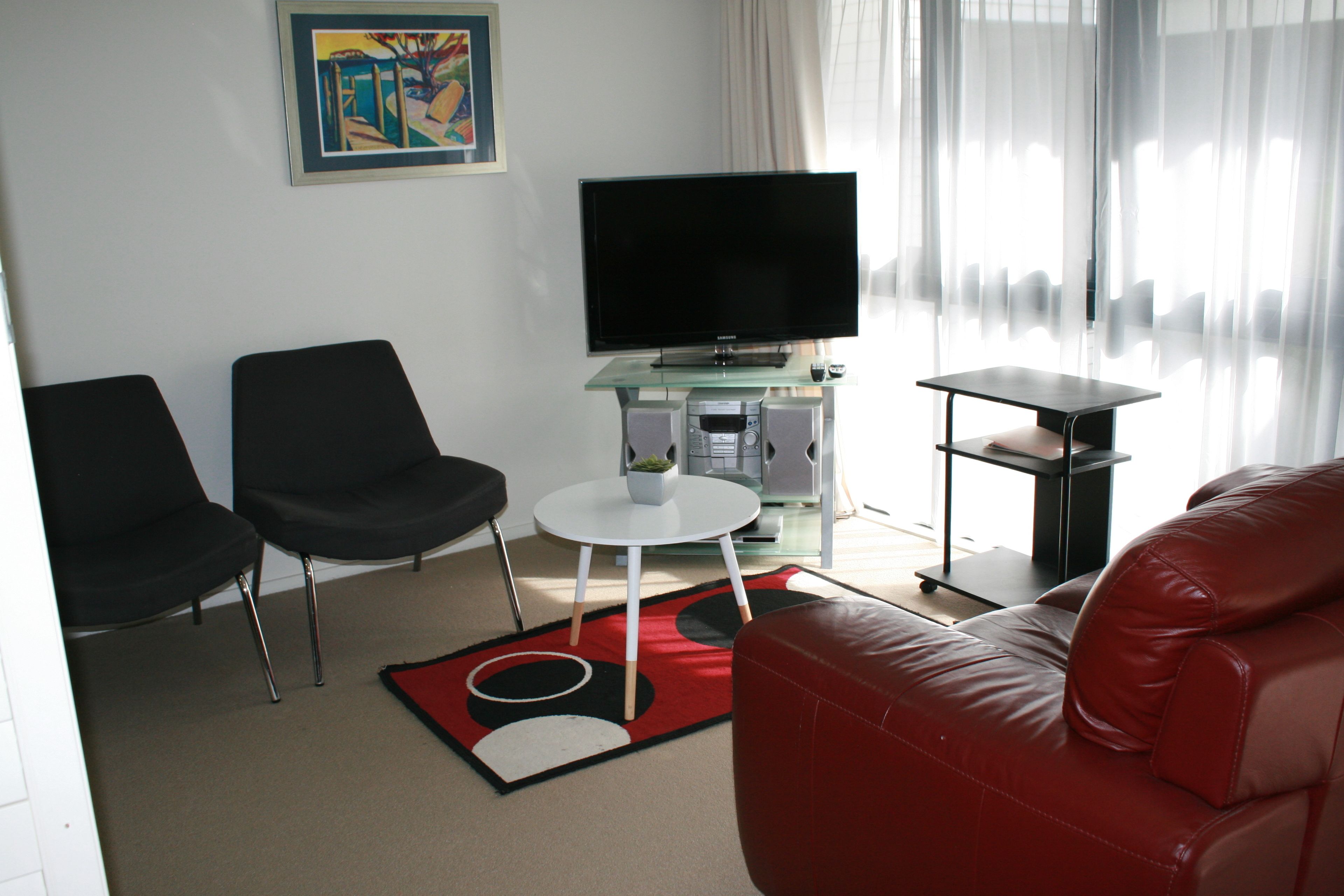 Un salon moderne avec un téléviseur à écran plat, deux fauteuils noirs, un canapé en cuir rouge et une table basse en verre.