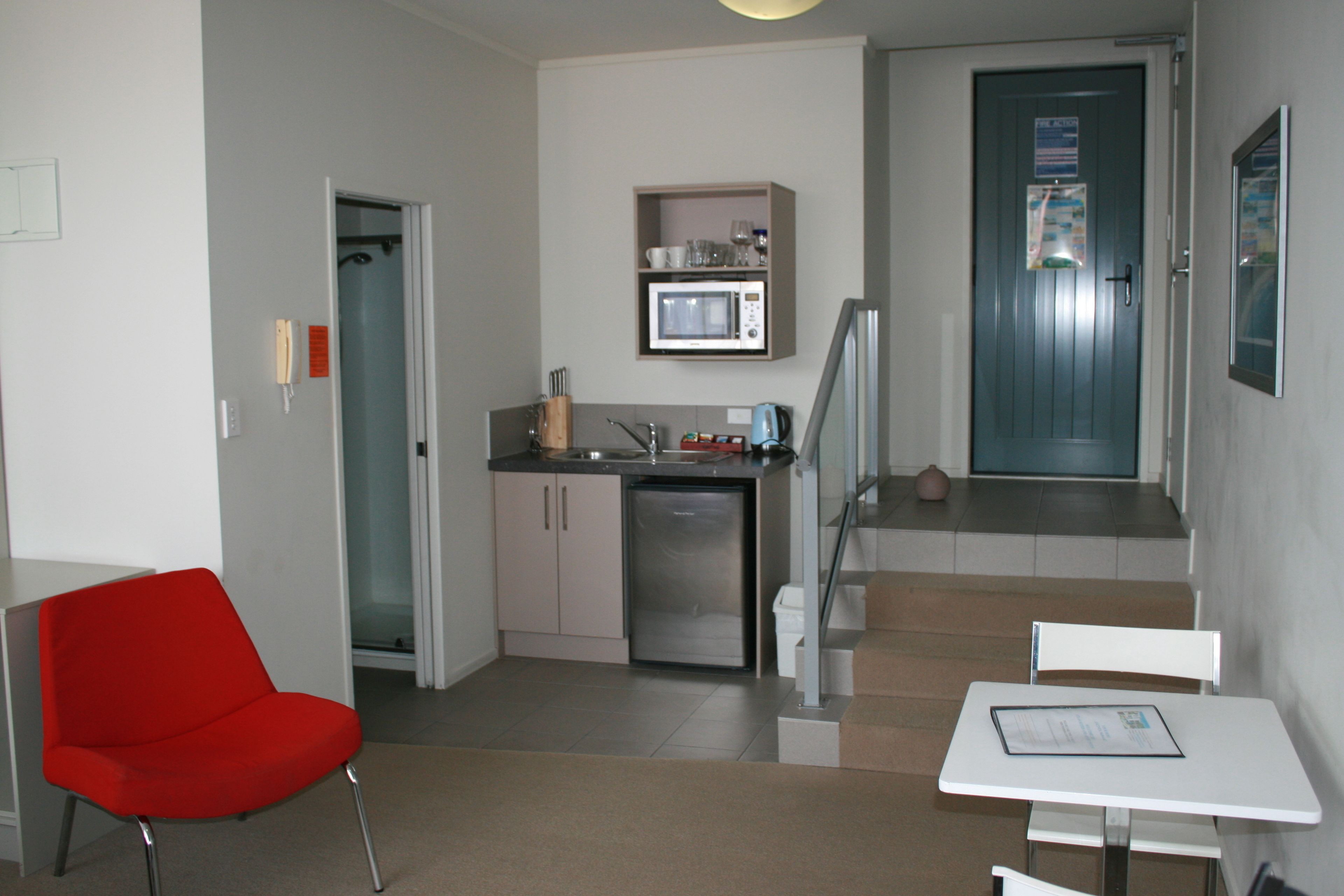 A compact living space with a kitchenette, a red chair, a white table, and a staircase leading to an upper level.