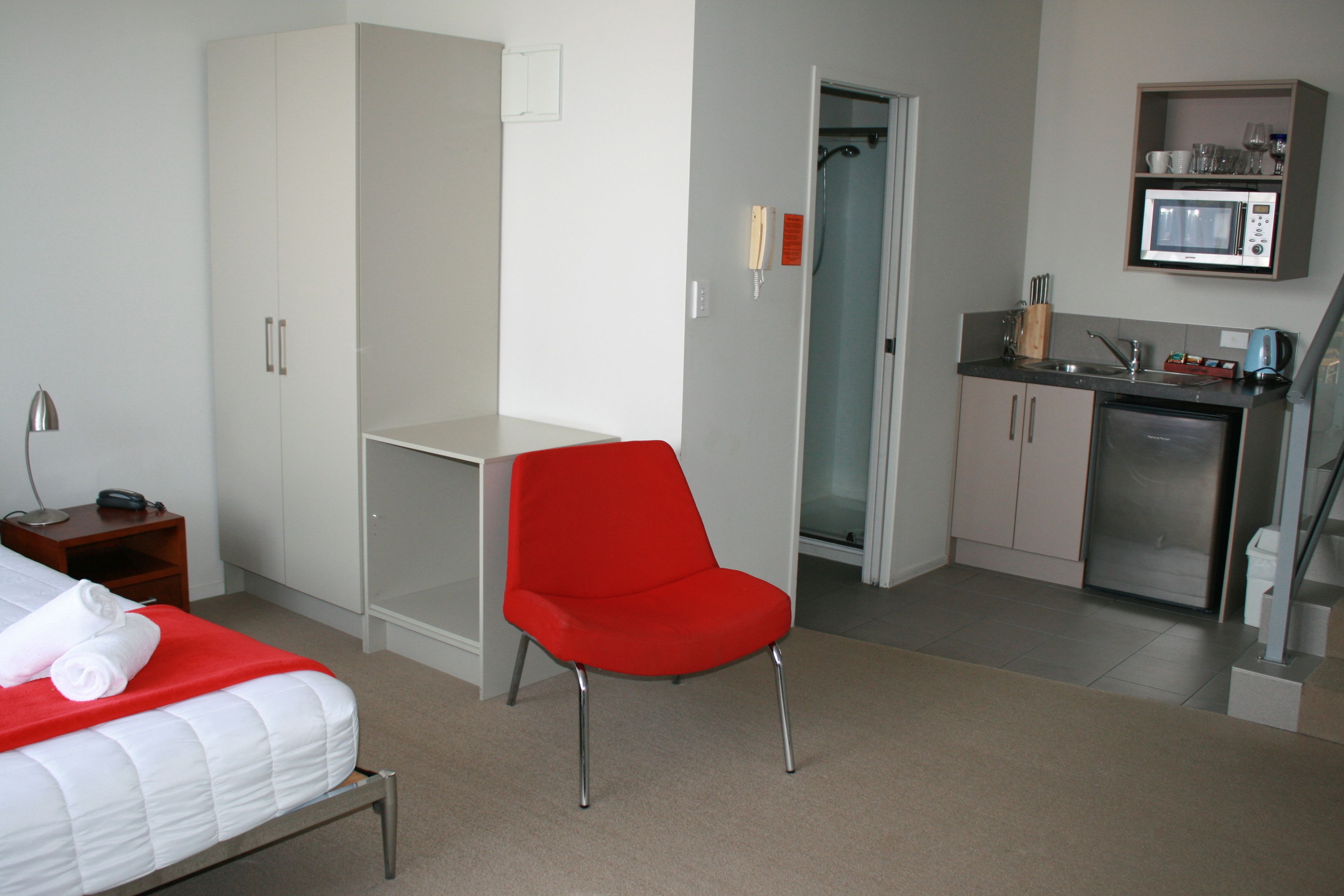 A compact hotel room with a bed, a red chair, a small desk, a kitchenette, and a shower area.