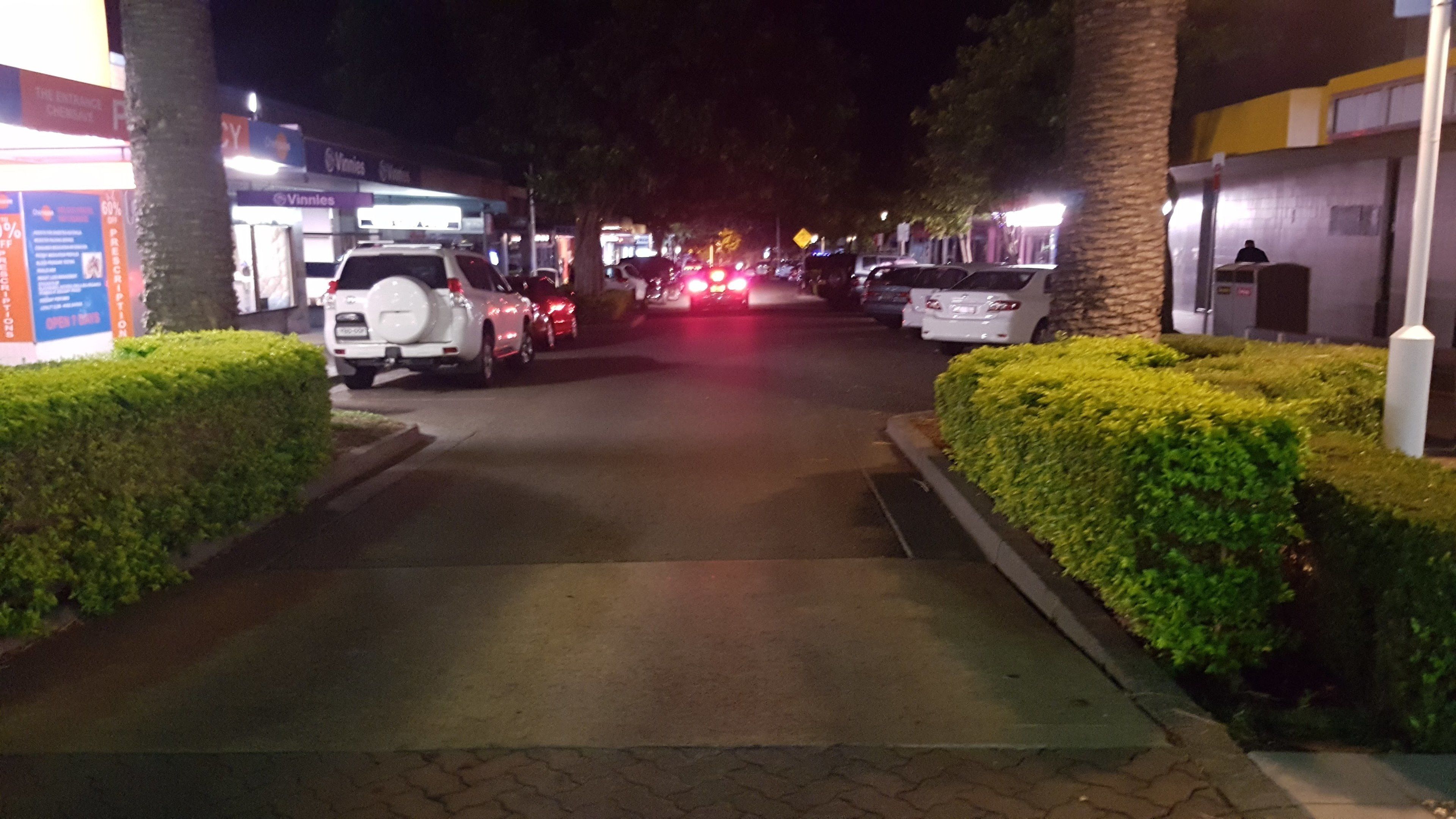 A nighttime street scene with parked cars, a sidewalk, and buildings with illuminated signs.