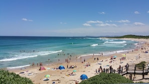 Una playa con gente nadando y tomando el sol, un muelle de madera y un cielo azul despejado.