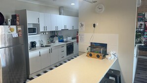 A modern kitchen with white cabinets, a stainless steel refrigerator, a microwave, and a sink. There is a clock on the wall and a poster with text. - The Entrance Backpackers - Hostel (The Entrance)
