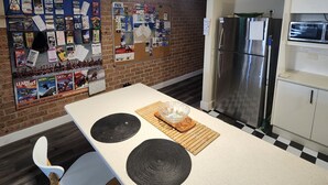 A kitchen with a white countertop, a stainless steel refrigerator, and a microwave. There is a wall with a bulletin board and a sign that reads "Nomads". - The Entrance Backpackers - Hostel (The Entrance)