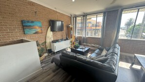 A living room with a brick wall, a black leather sofa, a flat-screen TV, and a window with a view of buildings. - The Entrance Backpackers - Hostel (The Entrance)