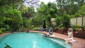 A pool surrounded by lush greenery, with a person sitting by the pool's edge. - Sunshine Valley Cottages (West Woombye)