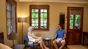 A room with a sofa, a small table with a lamp, a door, and a window with a view of greenery. - Sunshine Valley Cottages (West Woombye)