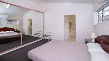 A bedroom with a large mirror, a bed with a wooden headboard, a nightstand with a lamp, and a small table with a chair.