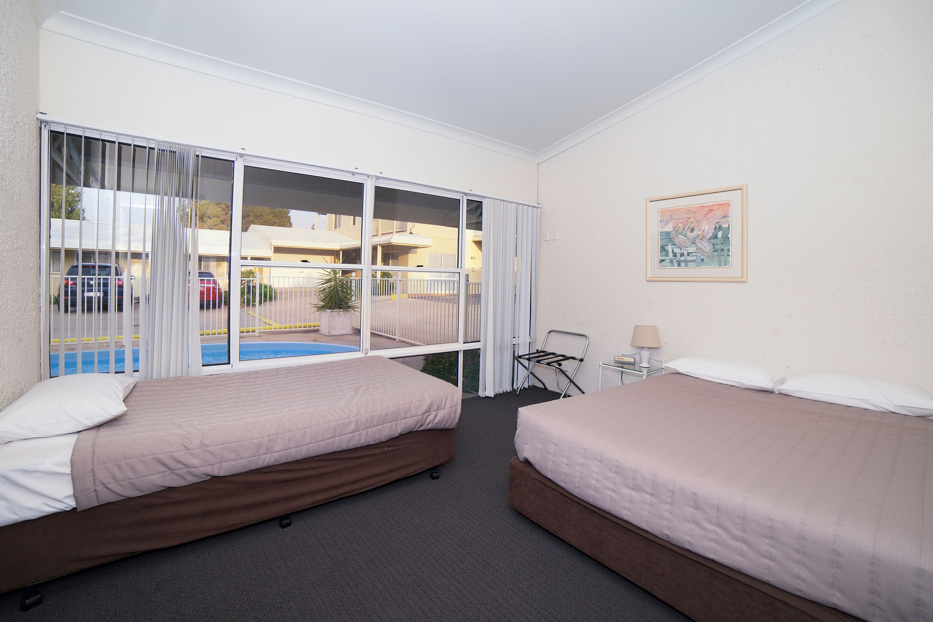 A hotel room with two beds, a window with curtains, a painting on the wall, and a view of a swimming pool and cars outside.