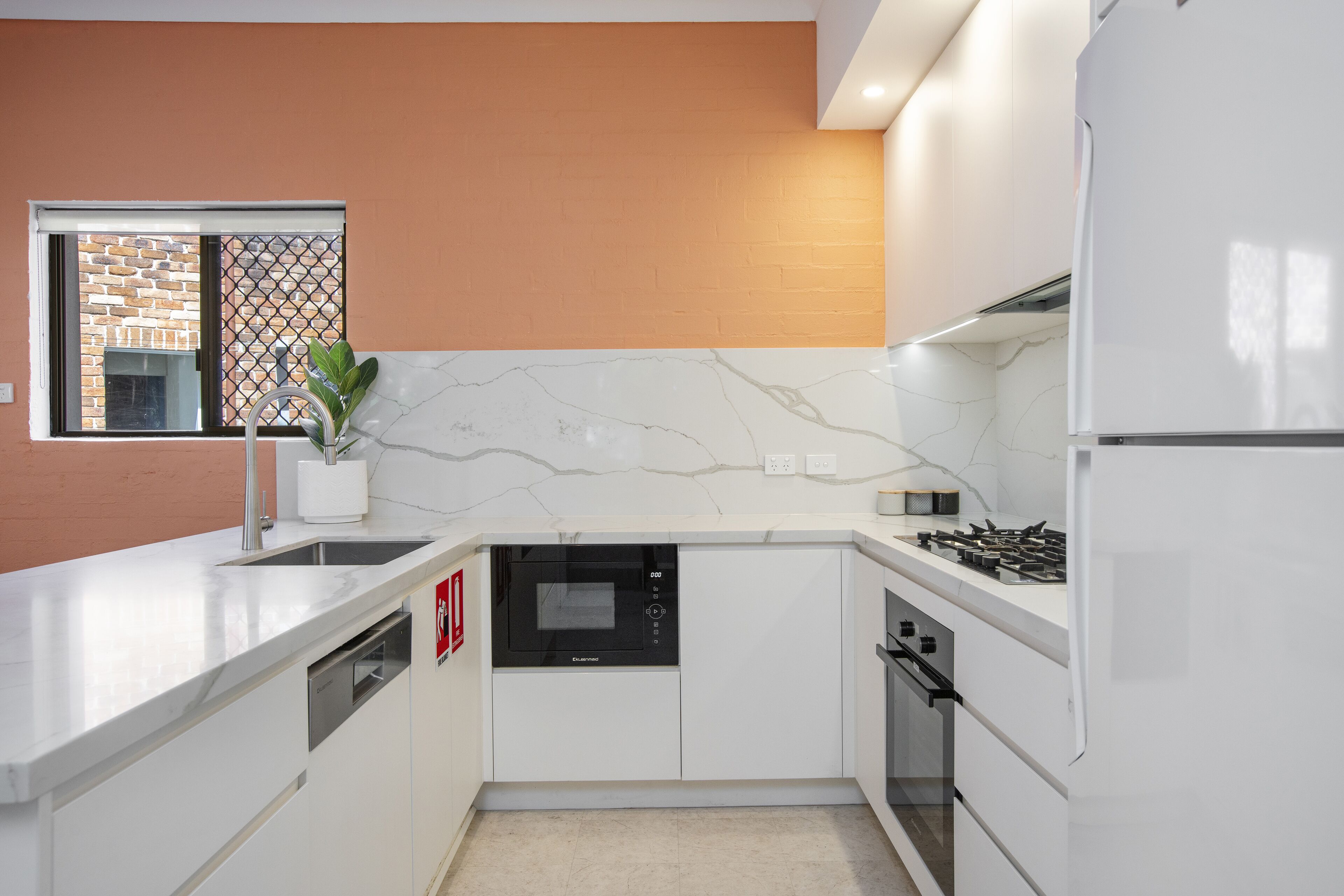 A modern kitchen with white cabinetry, a built-in dishwasher, a stove, and a window with a view of a brick wall.