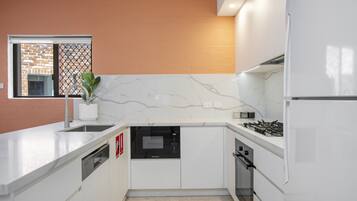 A modern kitchen with white cabinetry, a built-in dishwasher, a stove, and a window with a view of a brick wall.