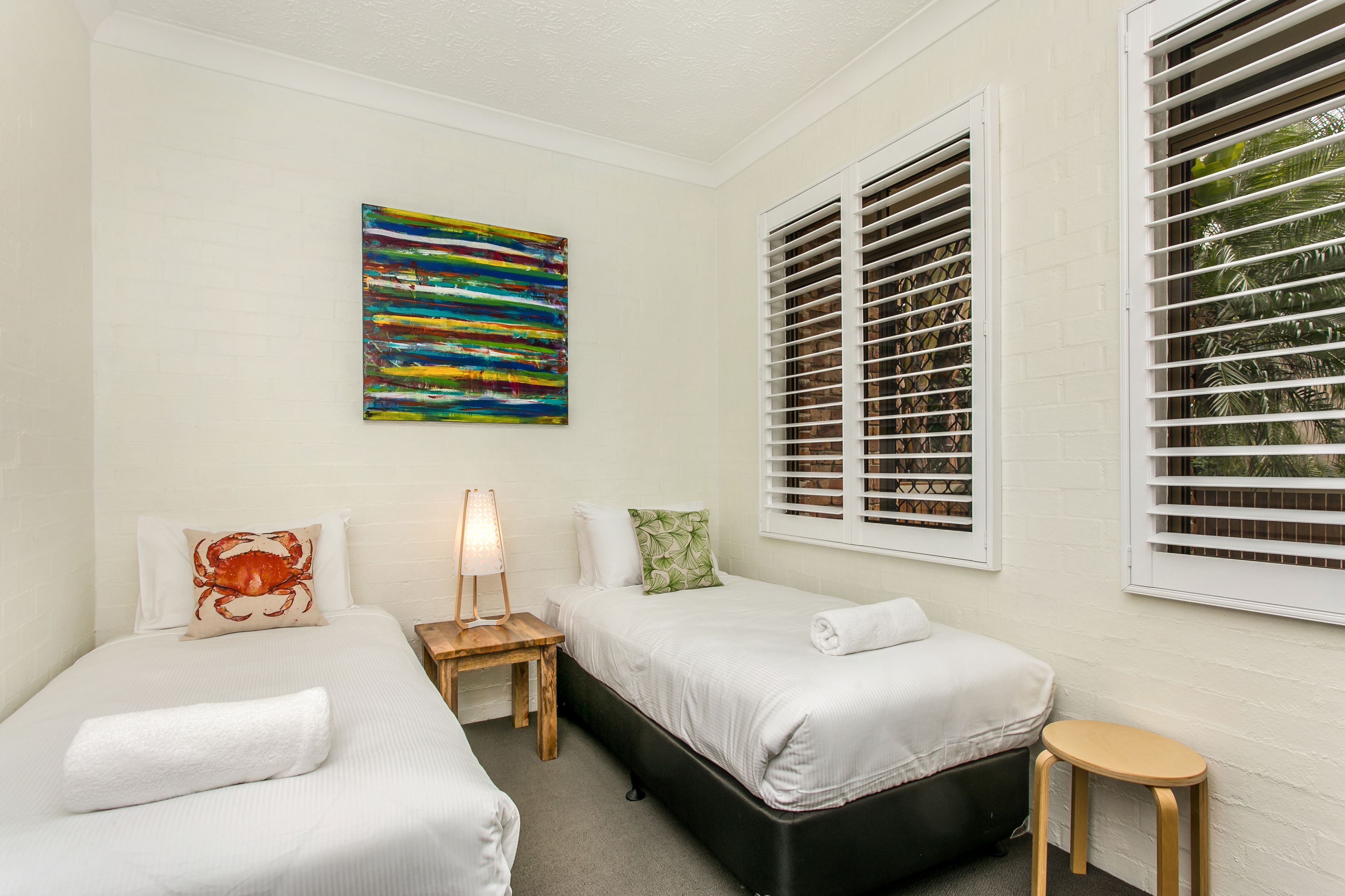 A hotel room with two beds, a painting, a lamp, and a window with blinds.