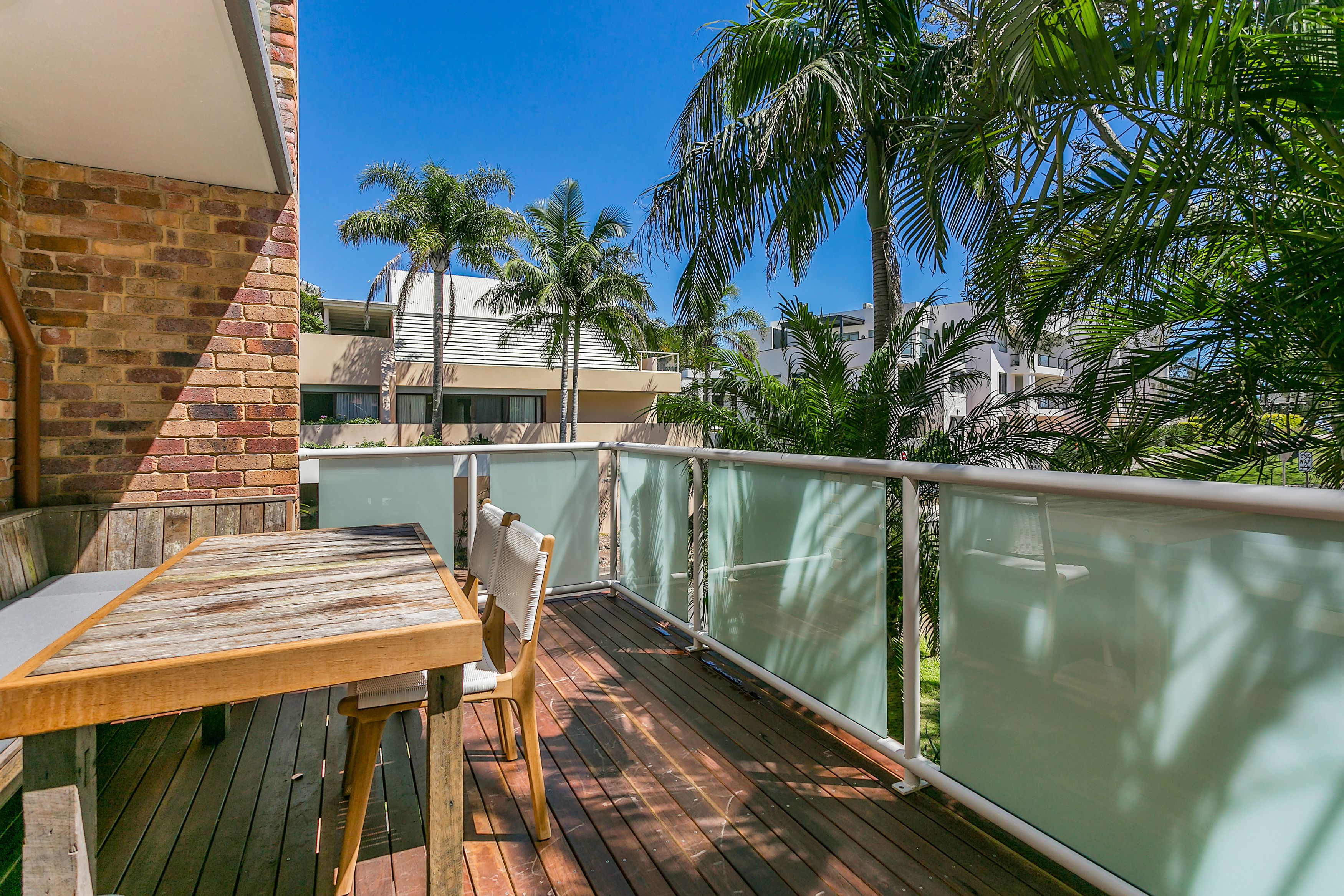 A wooden deck with a table and chairs, surrounded by a glass railing, overlooking a residential area with palm trees and houses.
