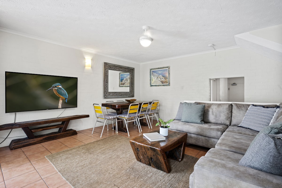 Ein Wohnzimmer mit Flachbildfernseher, einer Holzbank, einem grauen Sofa und einem Essbereich mit Stühlen und Tisch.
