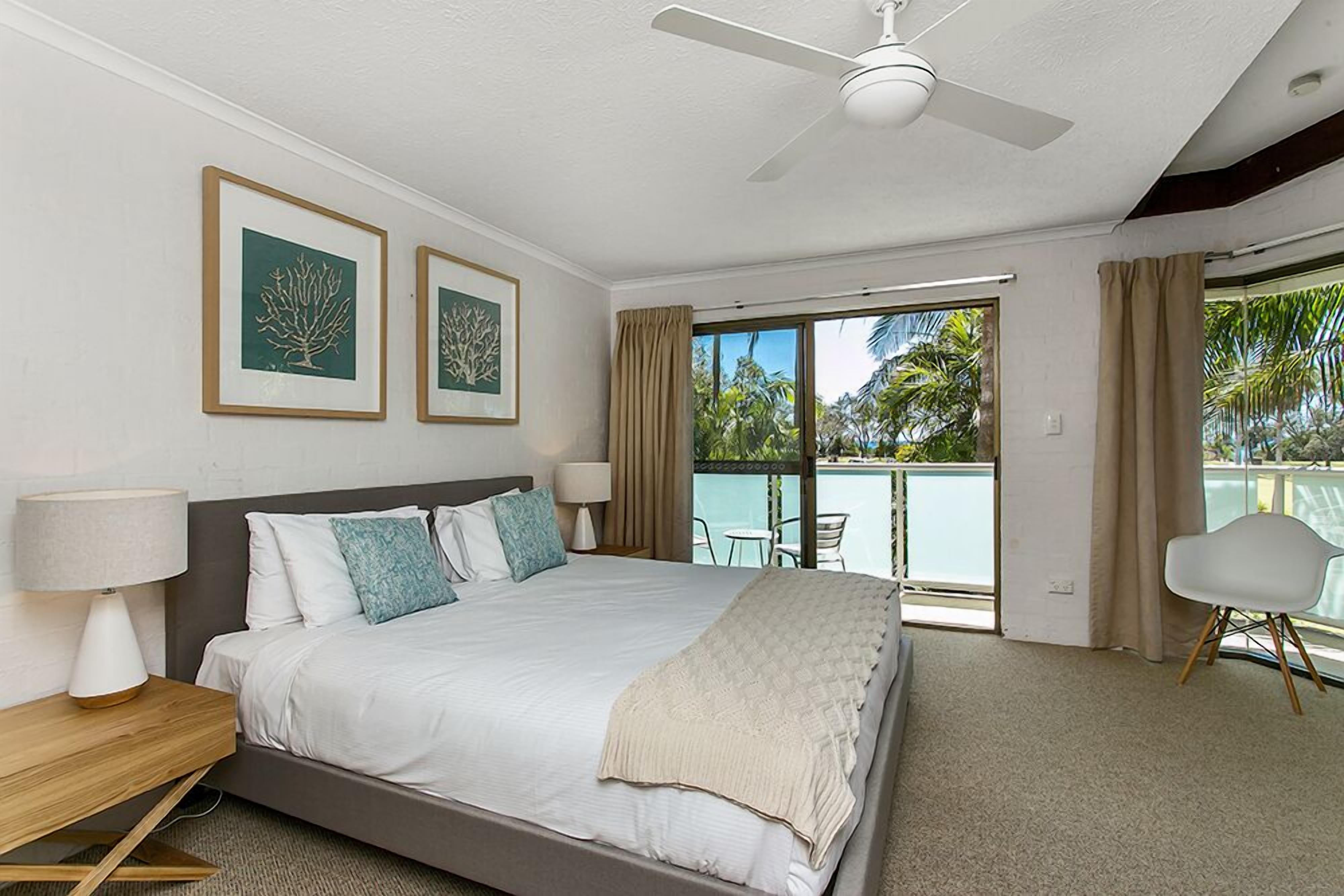 A hotel room with a bed, a ceiling fan, a window with a view of a pool and palm trees, and a chair by the window.