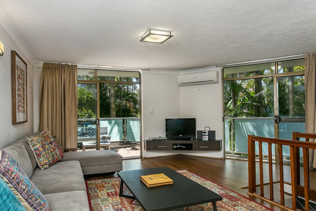 Ein Wohnzimmer mit einer Couch, einem Couchtisch, einem Fernseher und einem Balkon mit Blick auf Grünflächen.