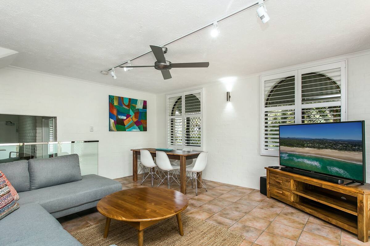 Ein Wohnzimmer mit grauem Sofa, einem Holztisch, einem Essbereich mit weißem Tisch und Stühlen, einem Deckenventilator und einem Fernseher auf einem Holzboden.
