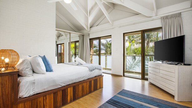 Une chambre moderne avec un grand lit, une tête de lit en bois, un téléviseur à écran plat et un balcon offrant une vue sur les palmiers.