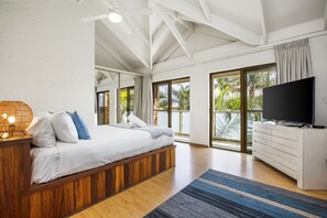 Un dormitorio moderno con una cama grande, cabecera de madera, televisor de pantalla plana y un balcón con vistas a palmeras.