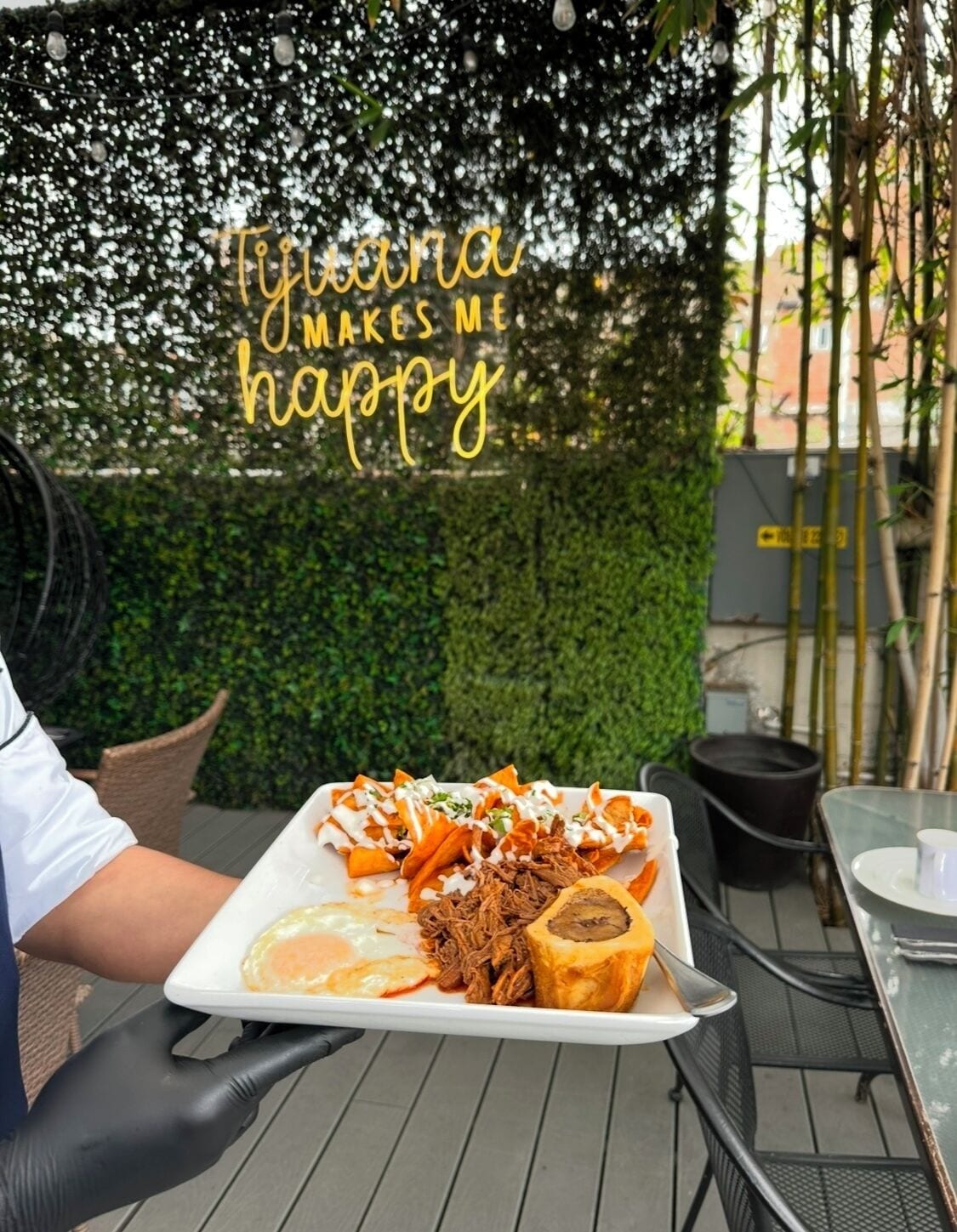 Una persona sosteniendo un plato de comida en una mesa al aire libre, con un muro decorativo al fondo.