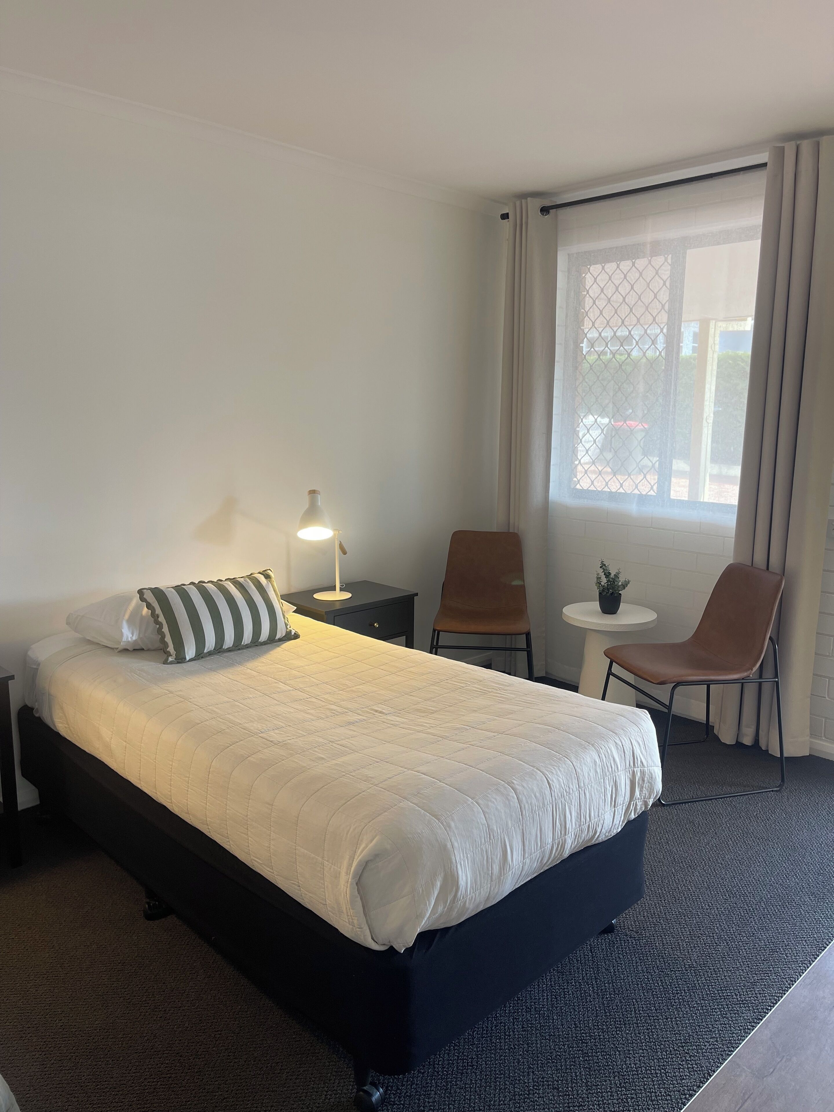 Habitación de hotel con cama, escritorio, silla, una pequeña mesa con planta y ventana con cortinas.