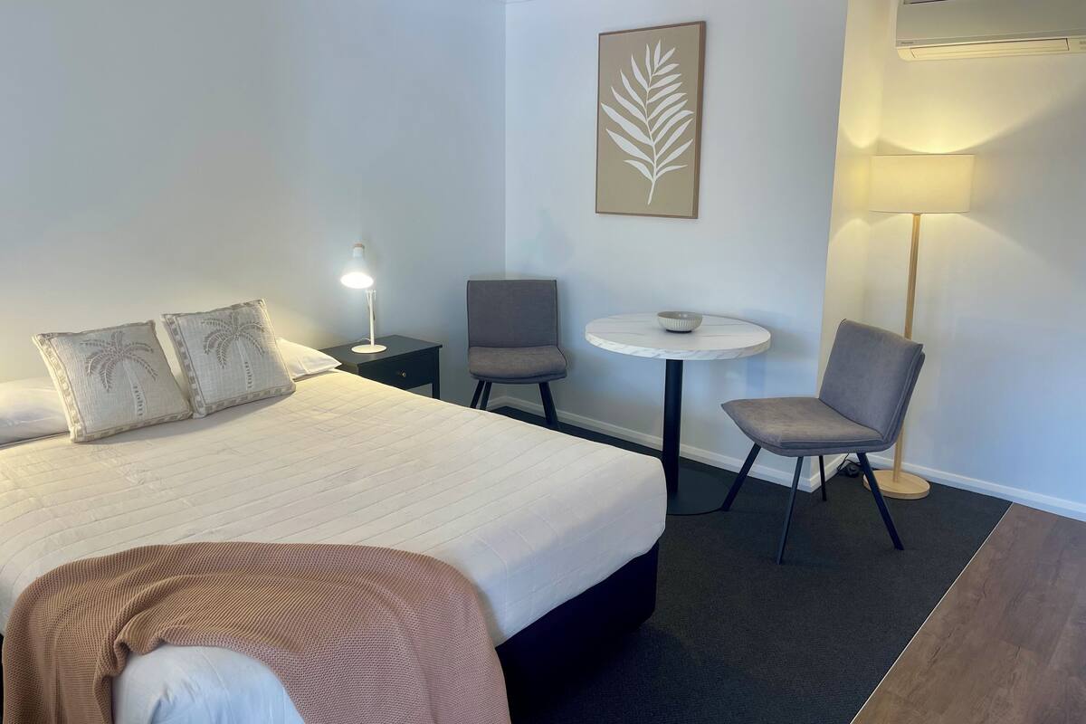 A hotel room with a bed, two chairs, a small table, a lamp, an air conditioner, and a wall-mounted artwork.