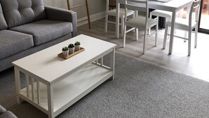 A modern living room with a grey sofa, a white coffee table, and a dining area with white chairs and a table.