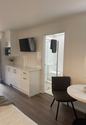 A modern kitchen with white cabinetry, a flat-screen TV, and a glass door leading to a bathroom. - Estelle Kramer Motor Inn (Armidale)