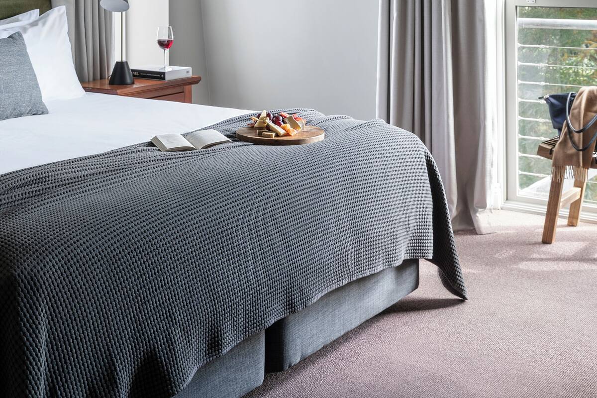 A neatly made bed with a grey and black checkered bedspread, a bedside table with a lamp and a plate of fruit, and a window with curtains.