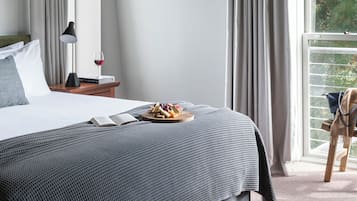 Una cama bien hecha con colcha a cuadros gris y negra, una mesita de noche con lámpara y una bandeja de fruta, y una ventana con cortinas.