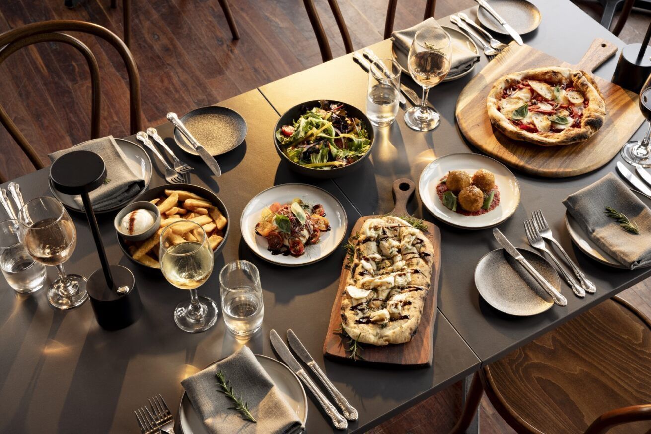Una mesa de comedor dispuesta con diversos platos, incluyendo pizza, ensalada y pan, junto con copas de vino y cubiertos.