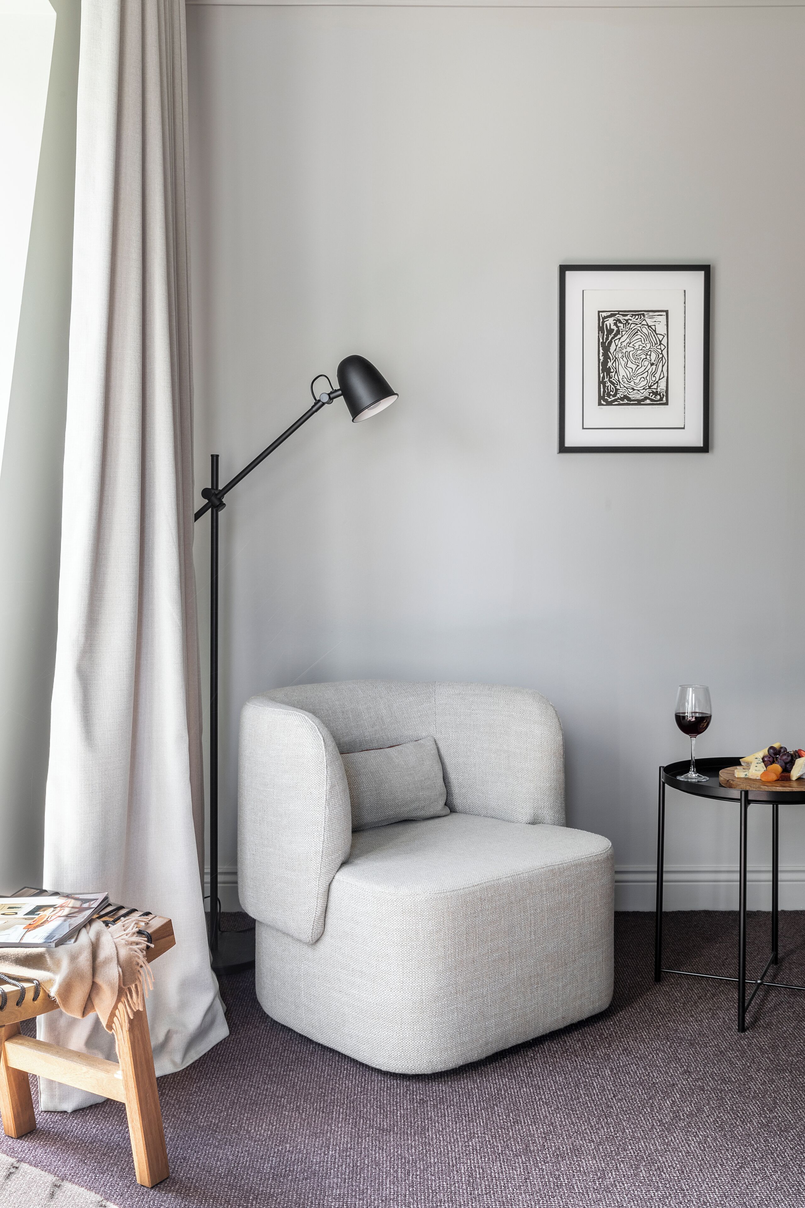 En moderne stue med en grå lænestol, en gulvlampe, et indrammet abstrakt kunstværk og et lille bord med et glas vin og ost.