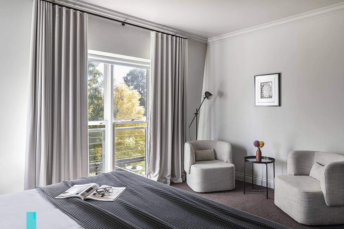 A modern hotel room with a large window, a bed with a striped bedspread, two armchairs, a small side table with a vase, and a framed picture on the wall.