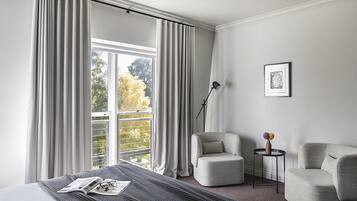 Habitación de hotel moderna con un ventanal grande, una cama con cubrecama a rayas, dos sillones, una mesita auxiliar con un florero y un cuadro en la pared.