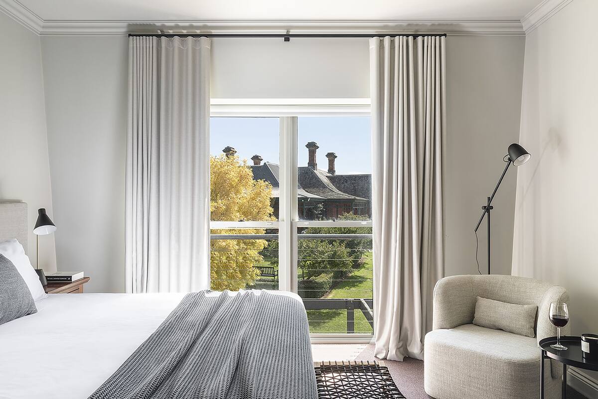 A bedroom with a large window, a bed, a chair, and a nightstand with a lamp.