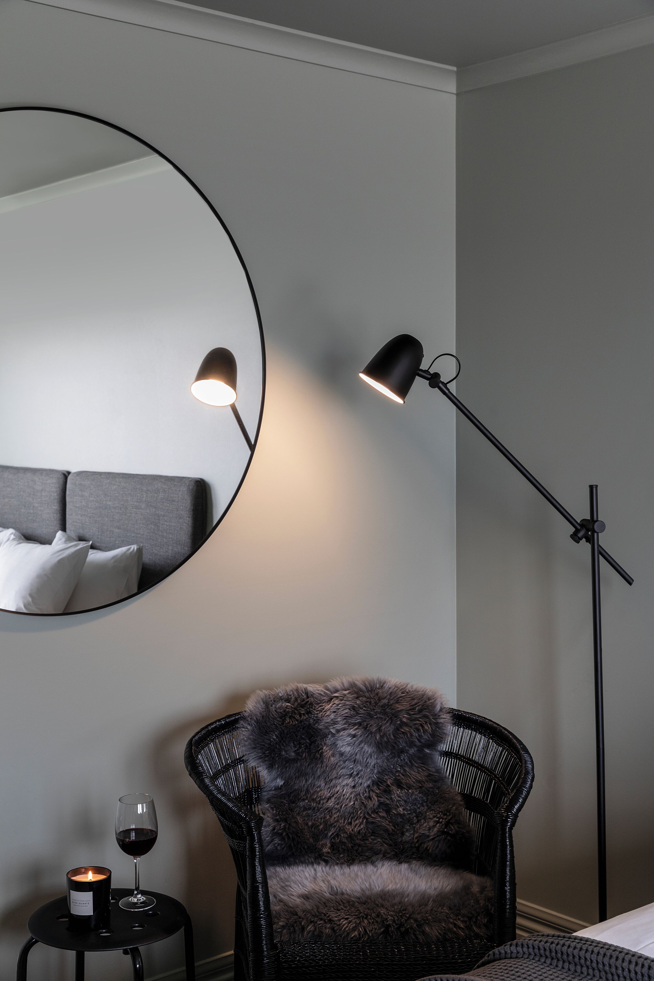 Una habitación moderna con un espejo redondo, una lámpara de pie, un sillón con una manta de piel, una mesita con una vela y una copa de vino.