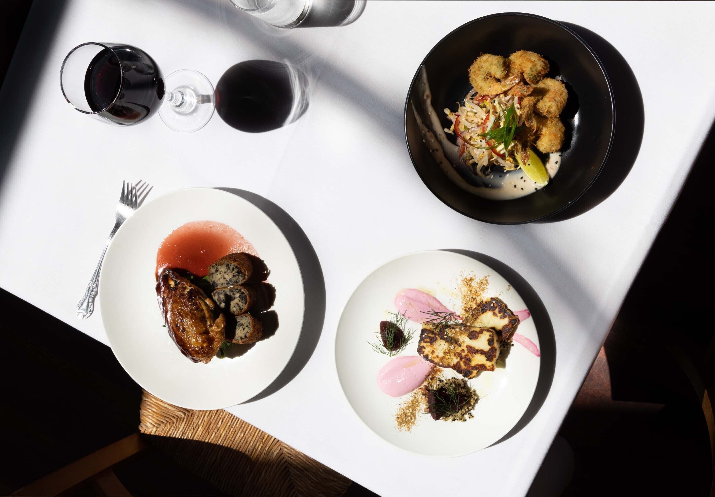Een tafel met drie borden eten, twee glazen rode wijn en een vork.