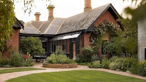 A traditional brick building with a gabled roof and chimneys, surrounded by a well-maintained garden with a paved pathway leading to a seating area. - Cleveland Estate (Lancefield)