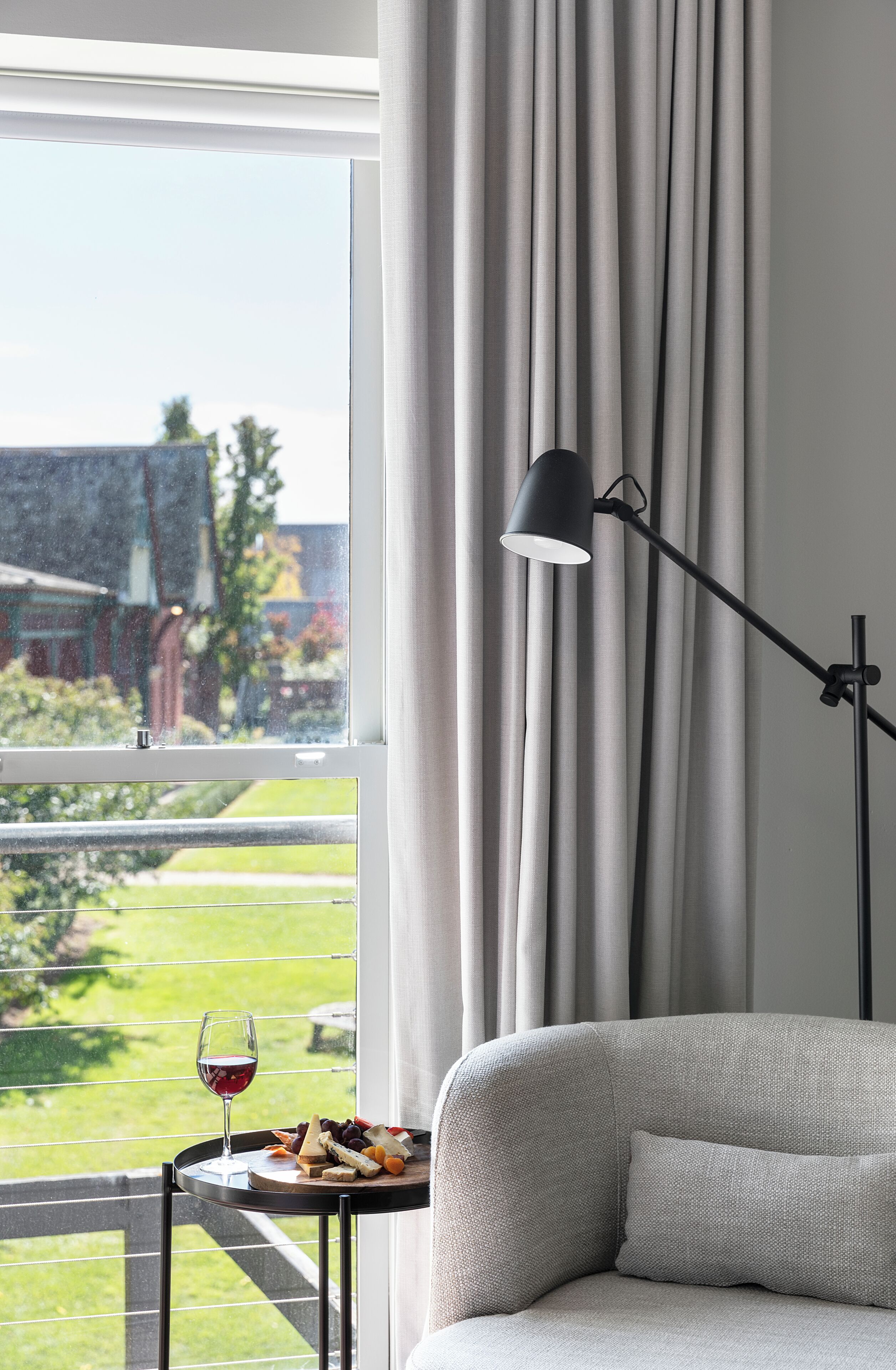 Een kamer met een bank, een kleine tafel met een glas rode wijn en een kaasplank, een vloerlamp en uitzicht op een groene tuin door een raam met gordijnen.
