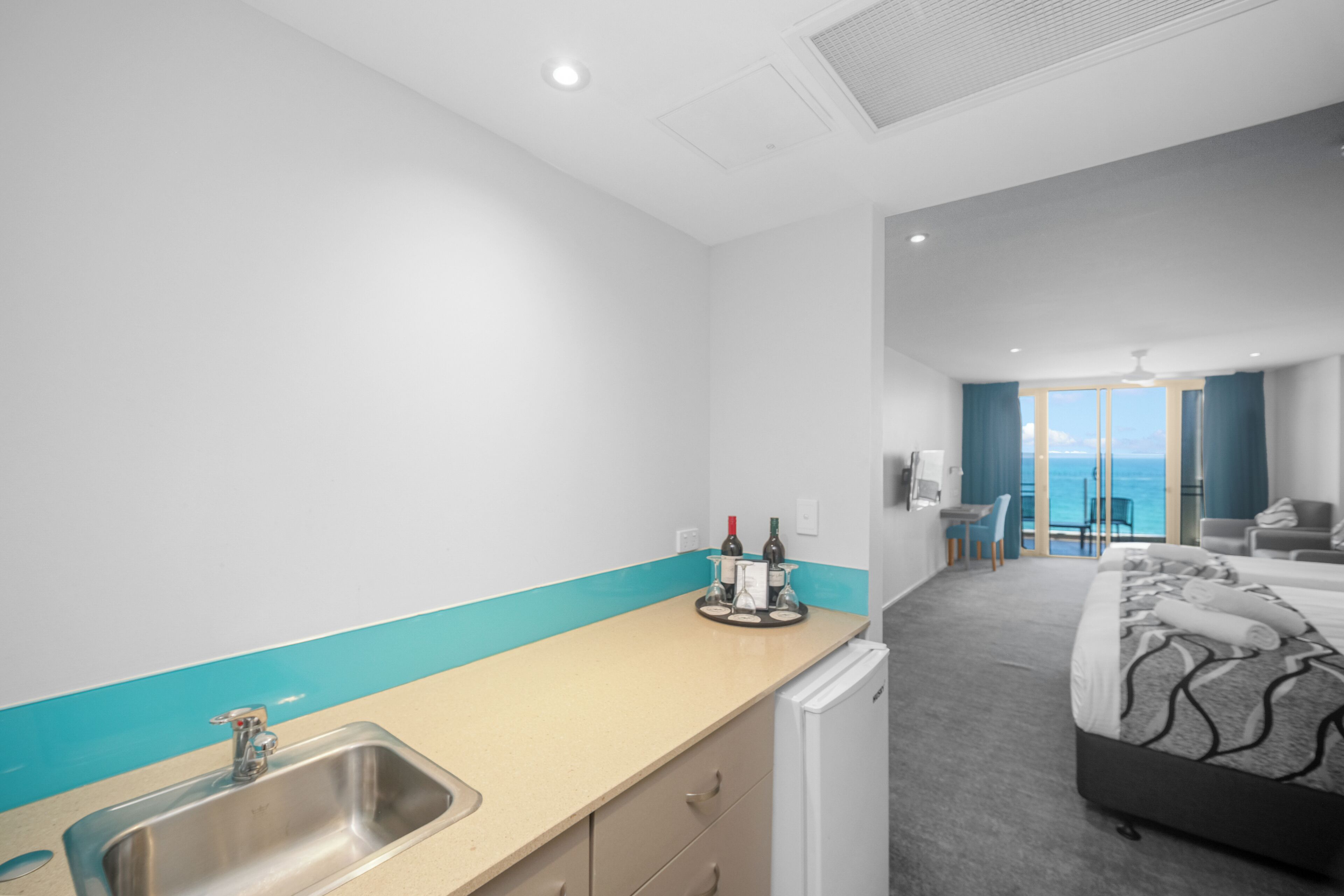 A modern hotel room with a kitchenette, a bed, a seating area, and a view of the sea.