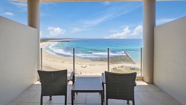 Un balcon avec deux fauteuils en osier et une table, donnant sur une plage où l’on peut admirer les vagues et un ciel dégagé.