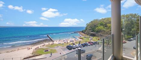 A beach view from a balcony with a clear sky, a parking area, and a road leading to the beach.