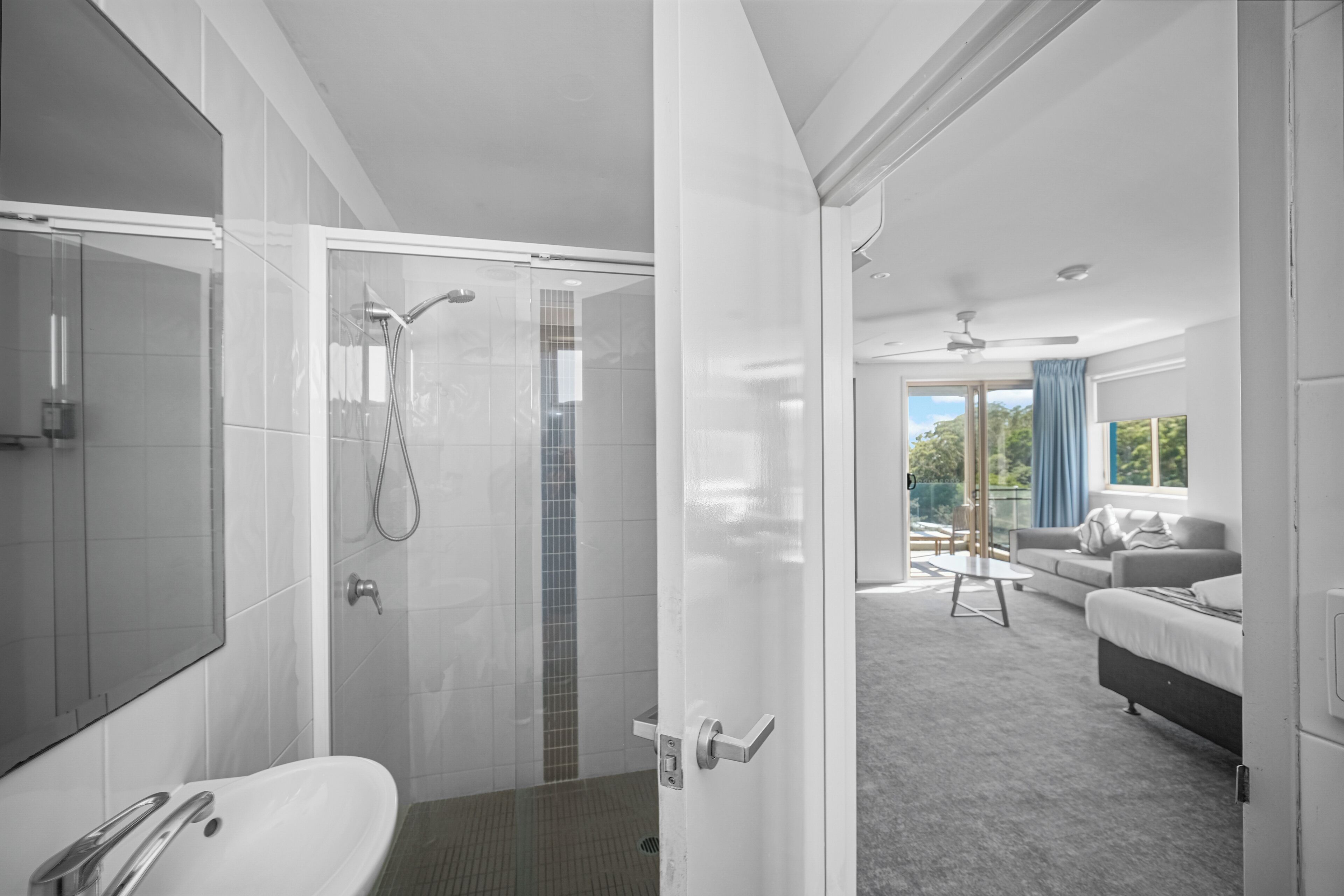 Et moderne badeværelse med bruser, vask og toilet. Der er en glasdør, der fører til en opholdsstue med sofa og bord.