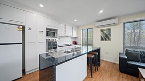 A modern kitchen with white cabinets, a granite countertop, and a built-in oven. There is a bar area with stools and a black leather couch in the adjacent living space. - Emerald Motel Apartments (Emerald)