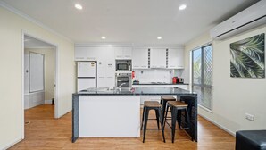 A modern kitchen with white cabinets, a central island with stools, and a large window with blinds. - Emerald Motel Apartments (Emerald)