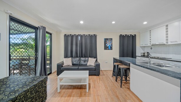 A compact living space with a sofa, a coffee table, a kitchen area with a countertop and sink, and a sliding glass door leading to an outdoor area. - Emerald Motel Apartments (Emerald)
