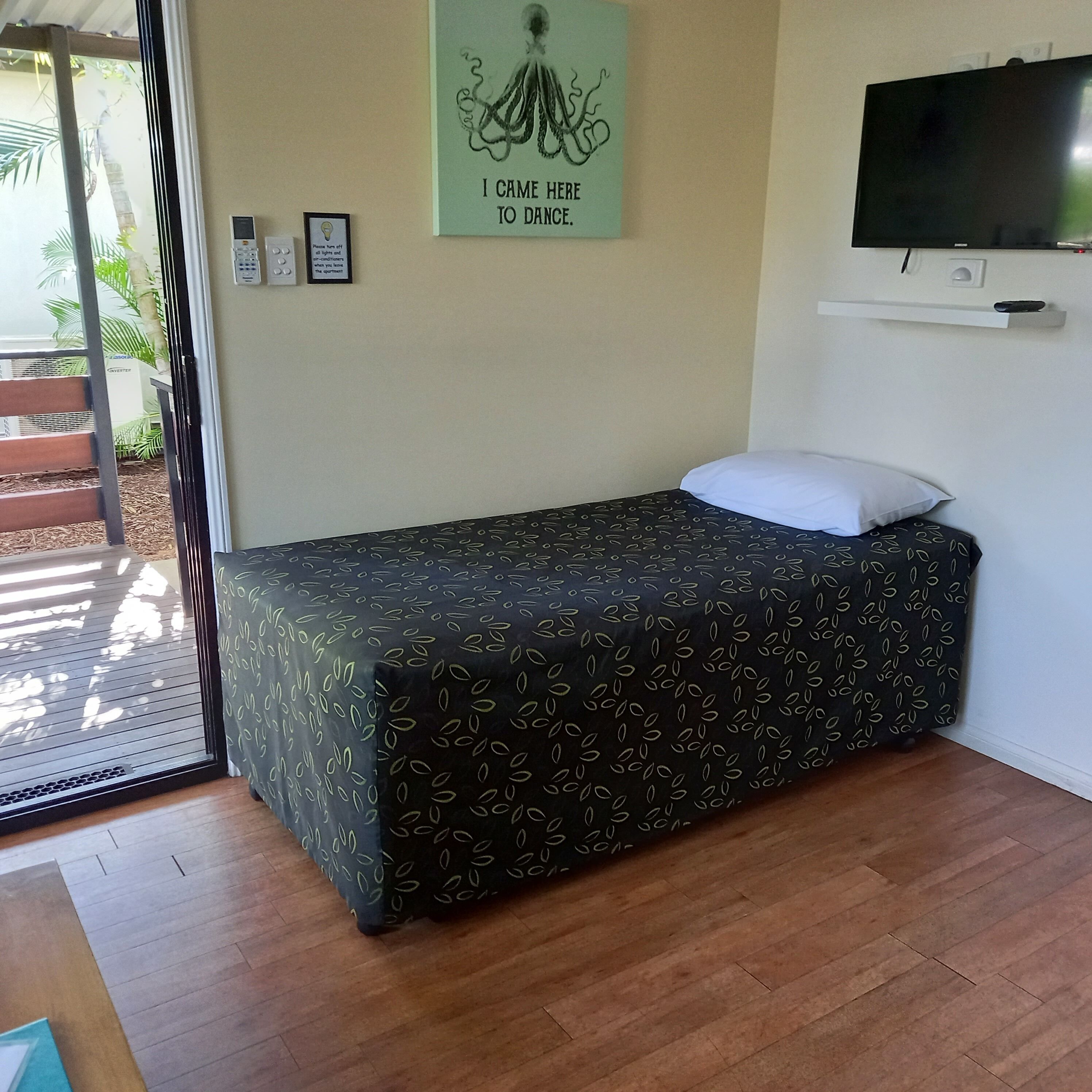 A single bed with a patterned bedspread, a wall-mounted TV, and a framed artwork with text.